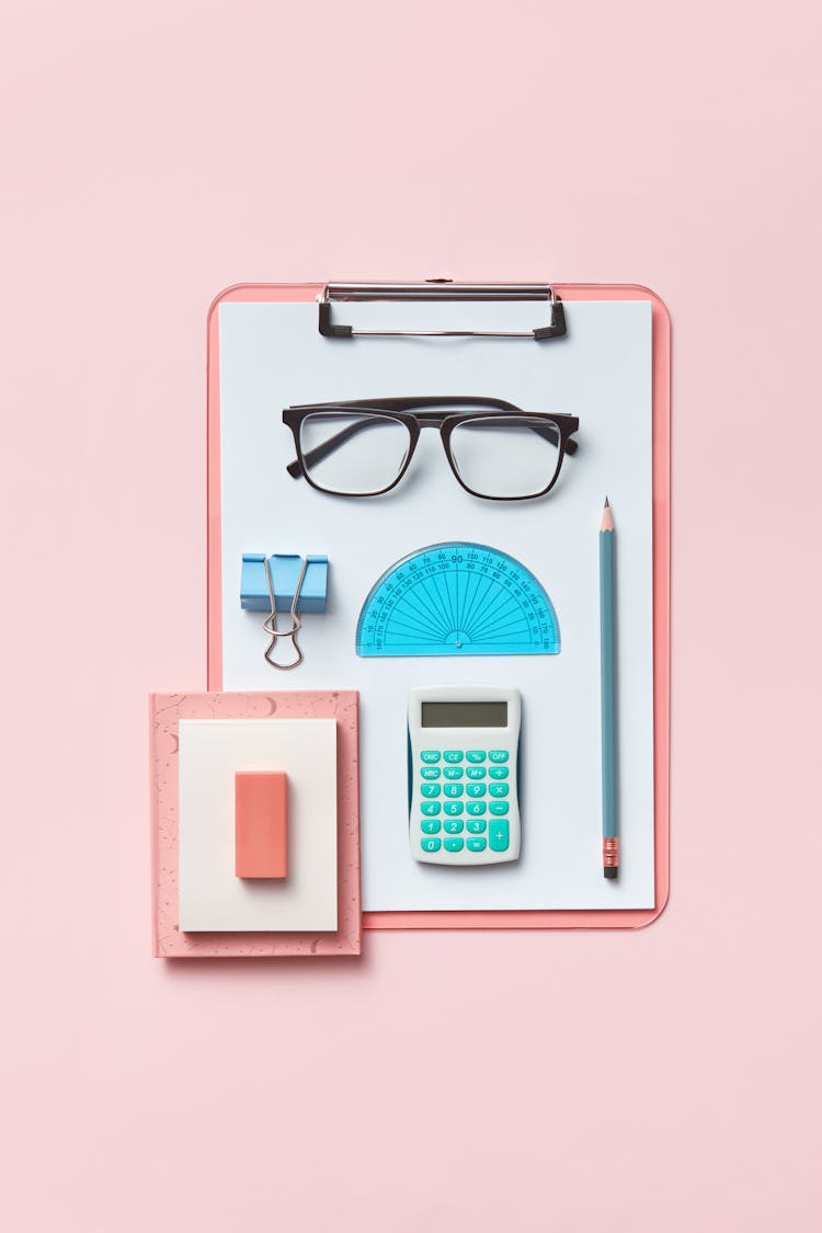 A Pencil, Calculator, Protractor, Clip Binder, And Glasses On A Pink Clipboard