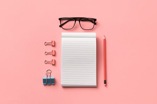 Free Flat lay of eyeglasses, notepad, pencil, and binder clips on a pink surface, ideal for creative workspace themes. Stock Photo