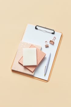 Flat lay of minimalist office supplies including clipboard, notepad, and pen on beige background.