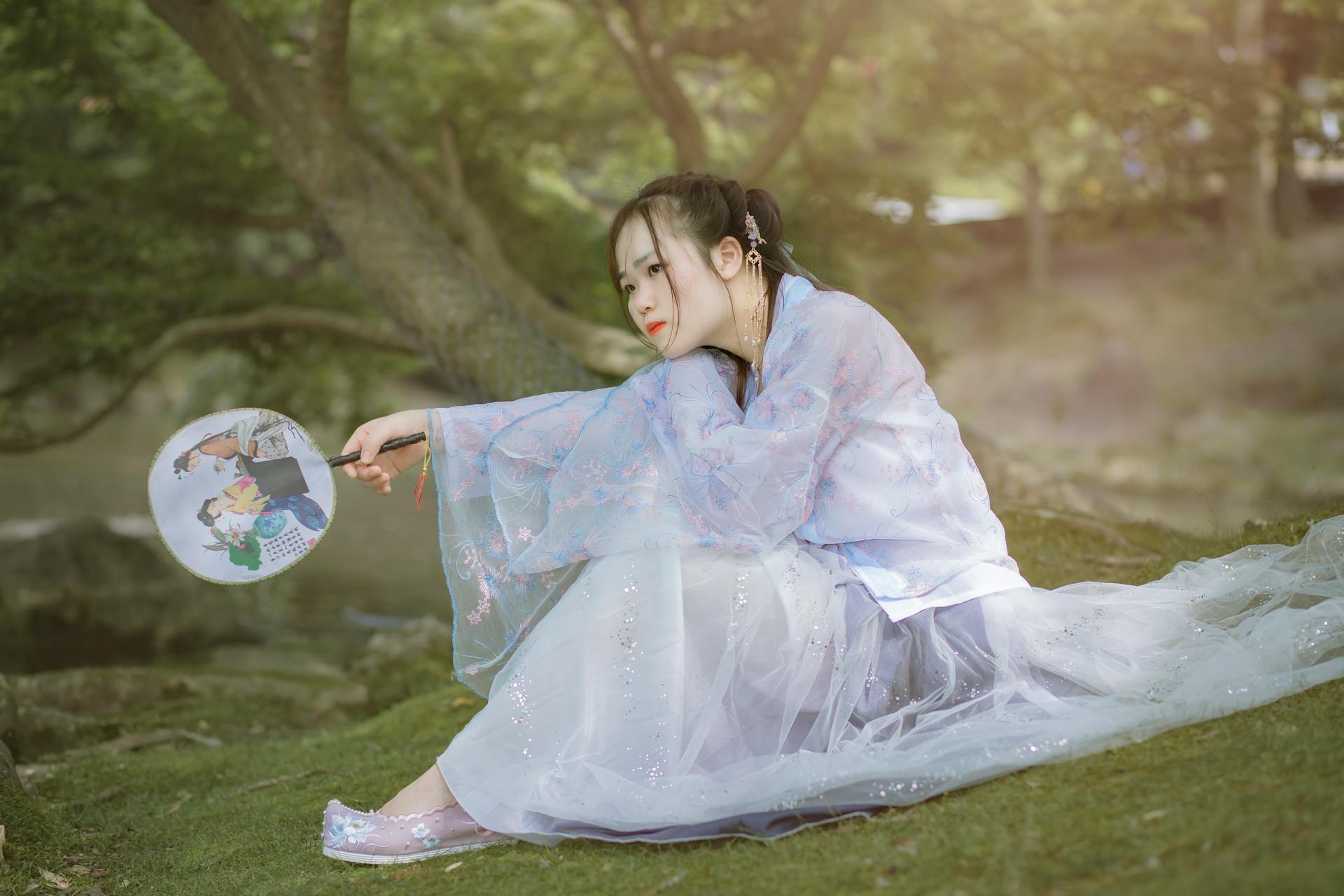 A woman in traditional Hanfu attire holding a fan while sitting in a serene outdoor setting.