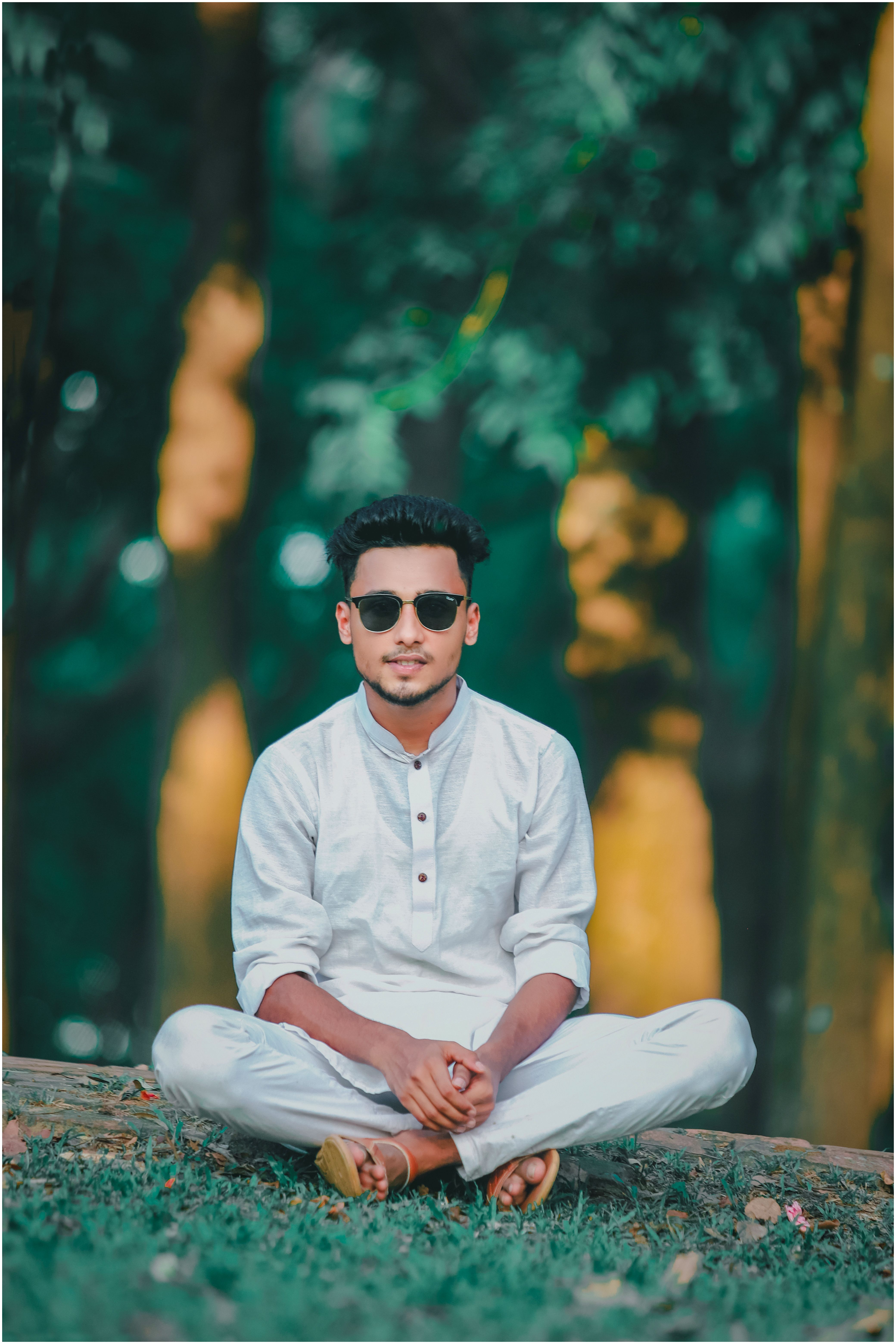 Young man in white outfit with sunglasses sitting outdoors in a forest setting.