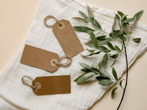 Free Three brown paper tags with twine on white fabric alongside green leaves. Eco-friendly design. Stock Photo