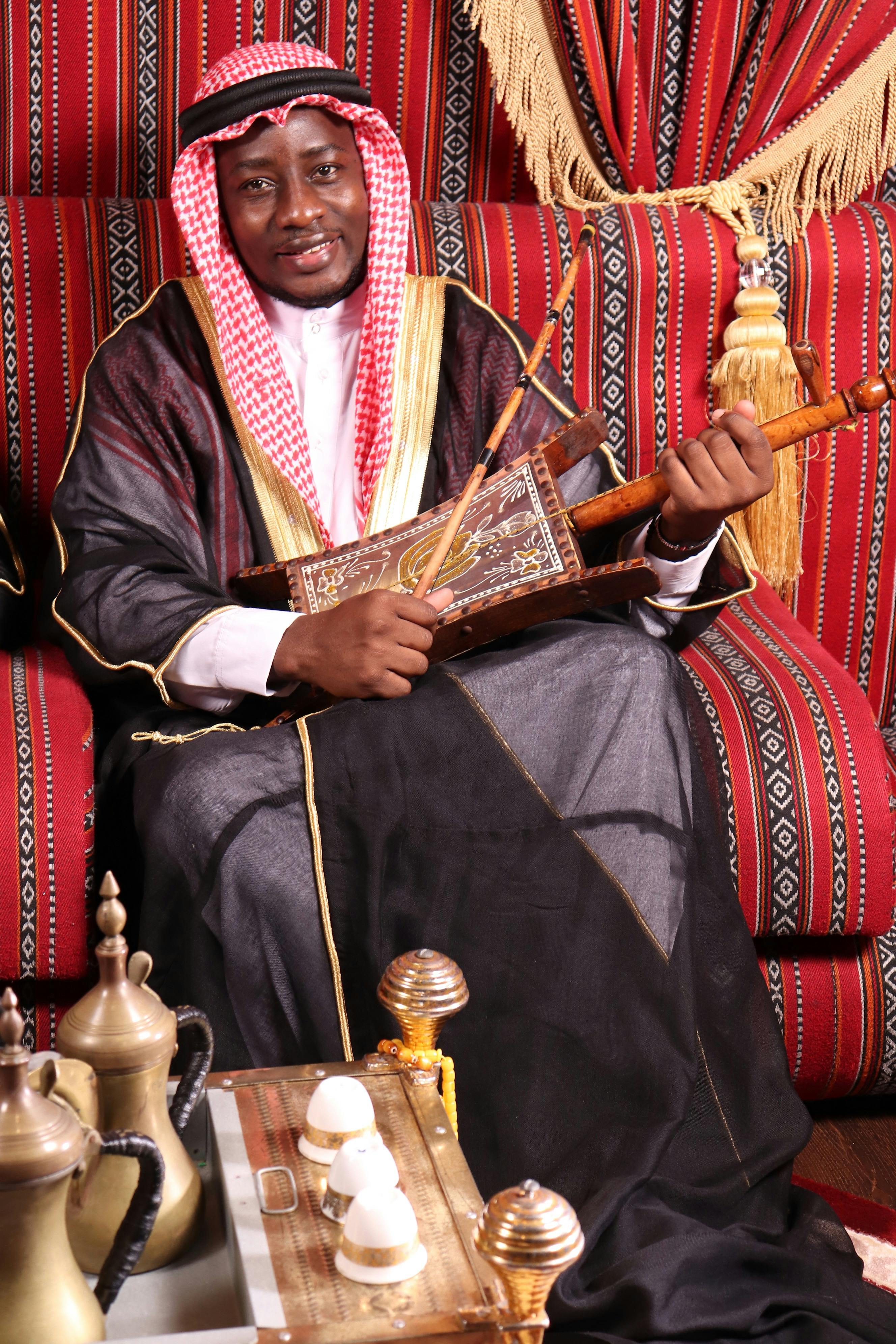 A man in traditional Middle Eastern attire playing a string instrument indoors on a patterned backdrop.