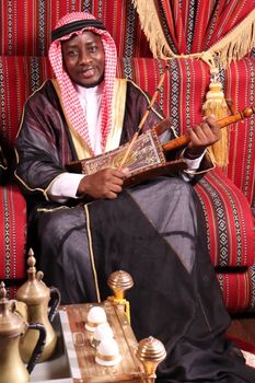 A man in traditional Middle Eastern attire playing a string instrument indoors on a patterned backdrop.