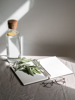 Simple composition with open notebook, eyeglasses, and water bottle on sunlit table.