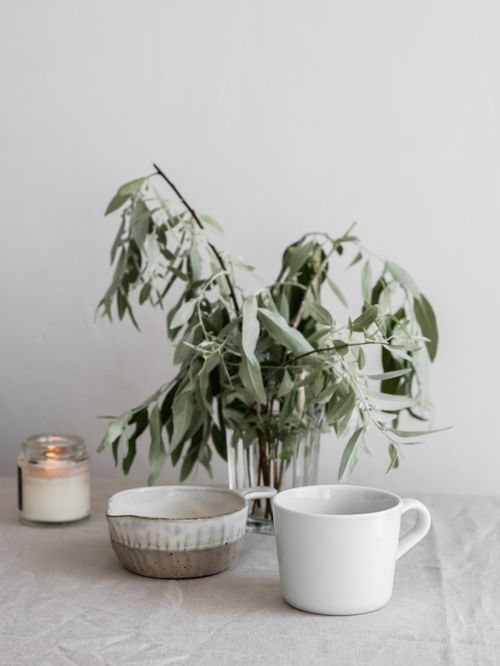 Free A serene composition of a ceramic mug, lighted candle, and leafy plant on a neutral surface. Stock Photo