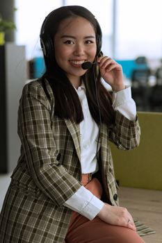Cheerful Asian woman in office setting wearing headset, offering customer service support.