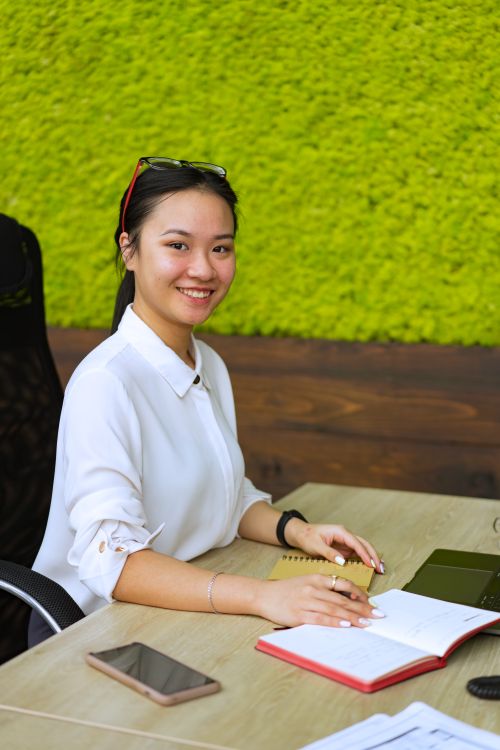 Free Asian woman smiling and working at a desk with green background in a modern office setting. Stock Photo