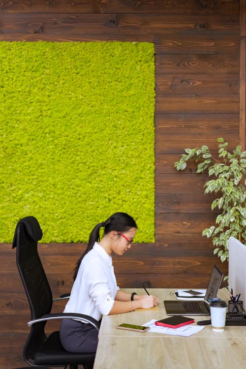 Free A focused woman writing at her desk in a stylish office, featuring a green wall accent and wooden decor. Stock Photo