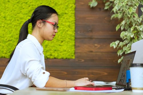 Free Asian woman working remotely on laptop in a stylish, modern office setting with green decor. Stock Photo