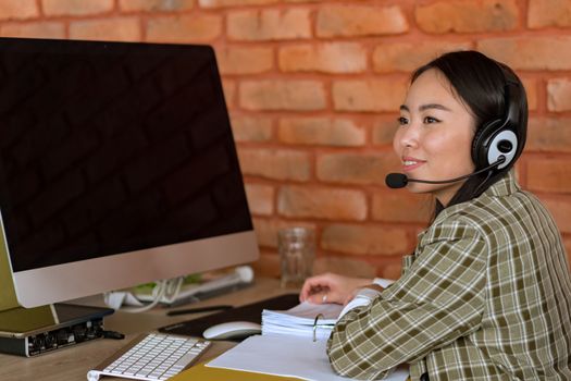 Asian woman in a plaid shirt working at a desk with a headset in a modern office.