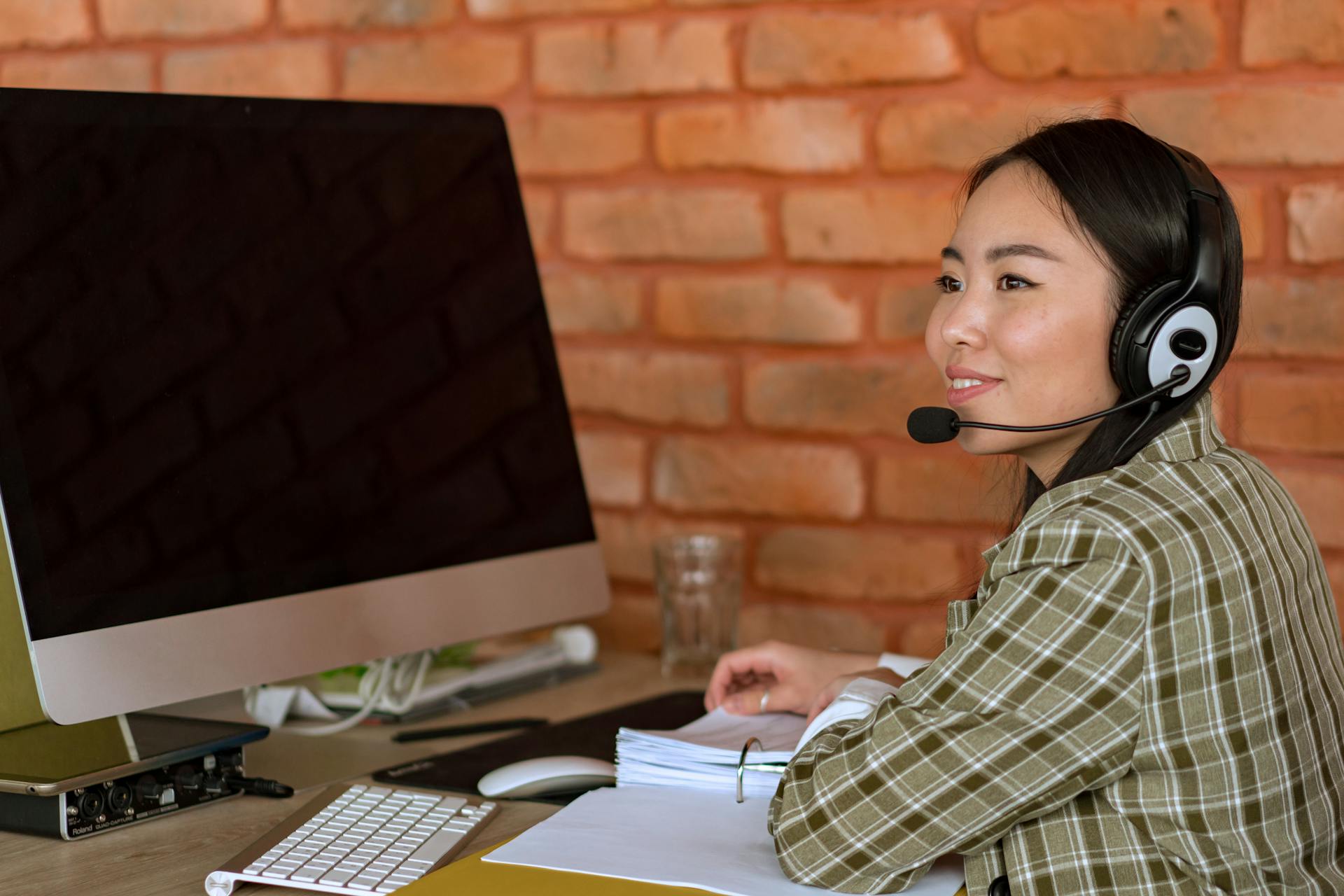 Asian woman in a plaid shirt working at a desk with a headset in a modern office.