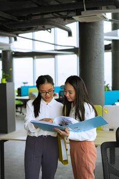 Two Asian women collaborating in a modern office setting with a binder.