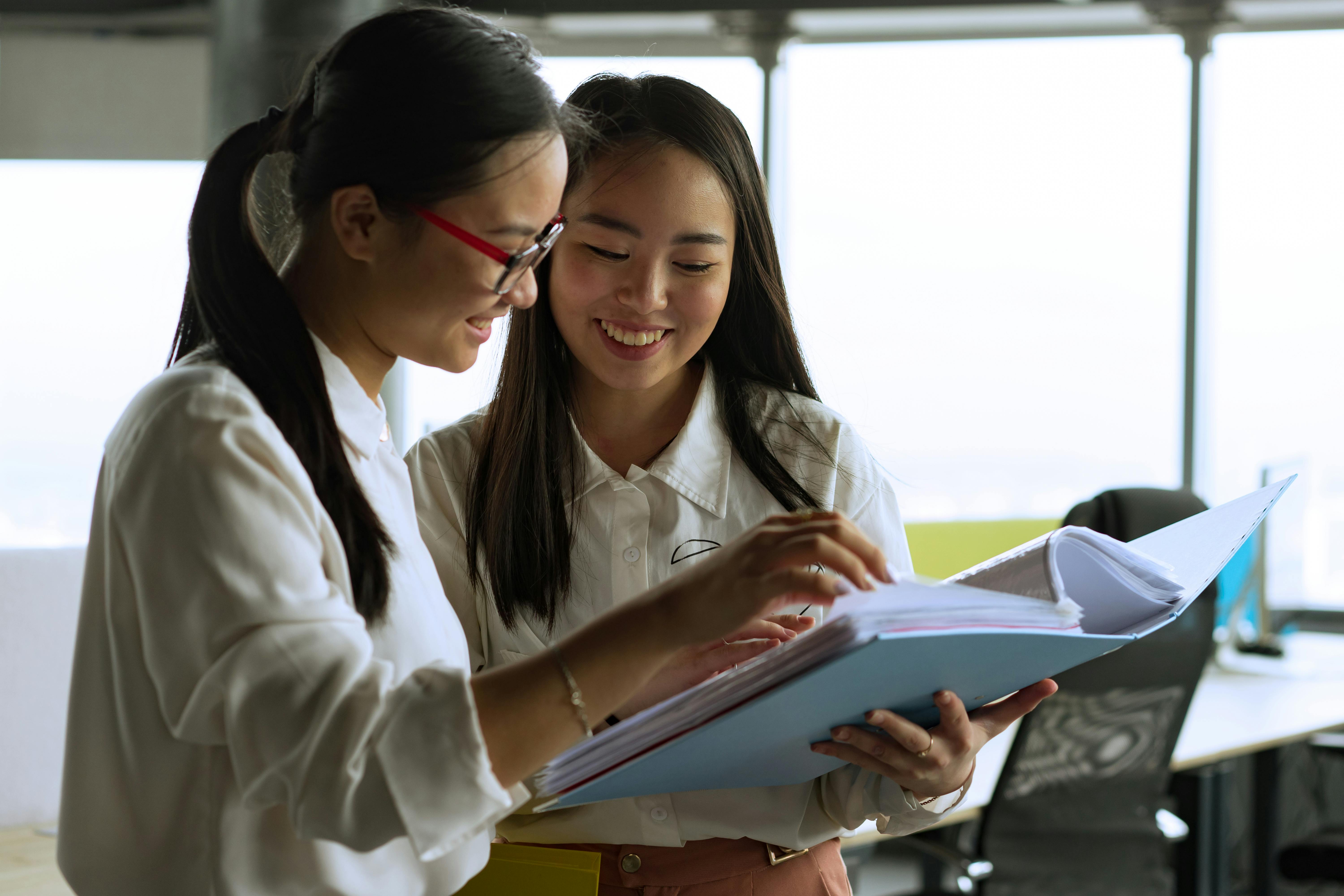 Two Asian businesswomen happily collaborating with a binder in a modern office space.