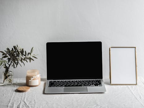 A sleek modern workspace featuring a laptop, candle, vase, and empty frame on a table.