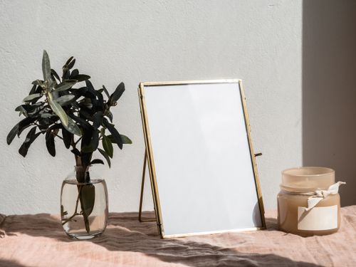 Free Minimalist scene with a blank frame, leafy plant in glass vase, and candle jar on a textured surface under soft daylight. Stock Photo