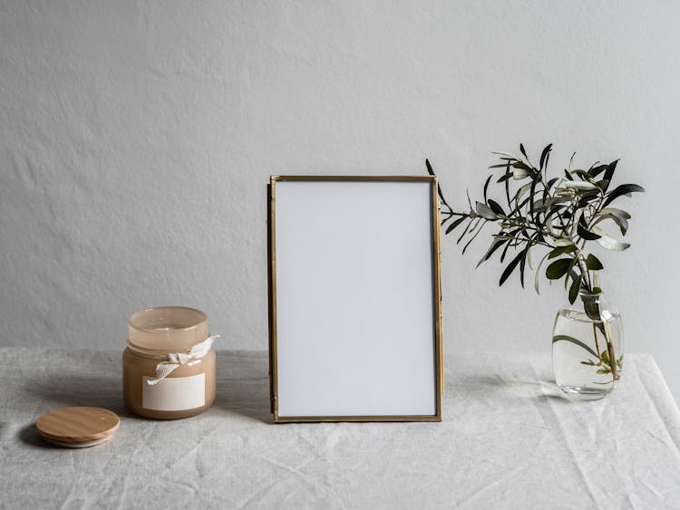 Empty Photo Frame Between A Glass Vase With Plant And Jar With Candle