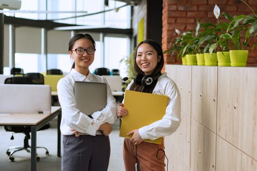 Free Two smiling Asian women in a modern office, holding documents and laptop, showcasing teamwork. Stock Photo