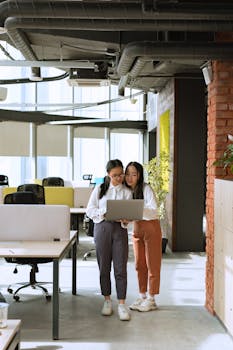 Two young women look at a laptop together in a bright, modern office, collaborating on a project.