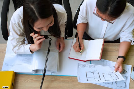 Two professional women collaborating in an office setting, using phone and taking notes.