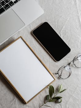 Flat lay of a workspace with a laptop, smartphone, glasses, and plant on a linen surface.