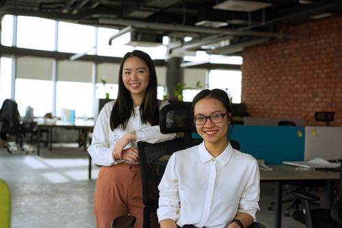 Free Two professional women smiling at camera in a modern office environment, showcasing teamwork. Stock Photo