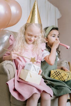 Two young girls enjoy a festive birthday celebration, surrounded by balloons and gifts indoors.