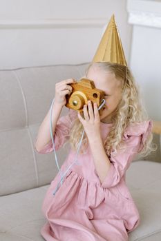 A young girl in a party hat plays with a wooden camera indoors.