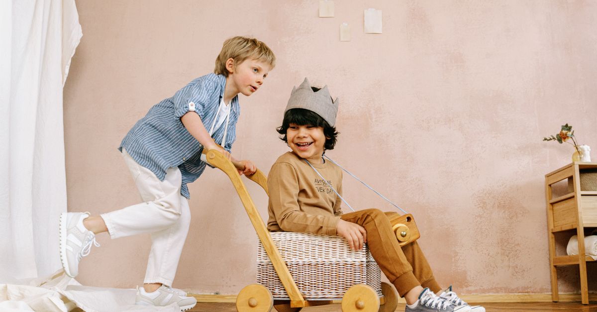 Two children playing with a handmade wooden toy cart indoors, smiling and enjoying themselves.