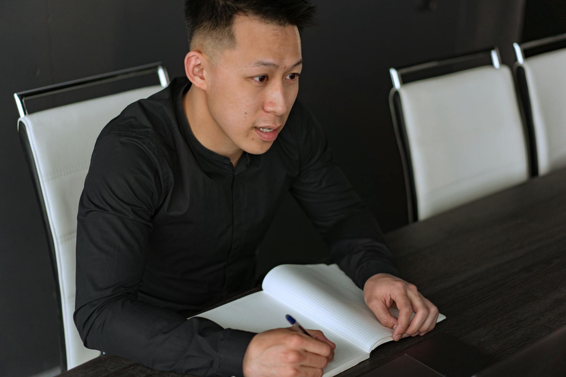 Asian man in black shirt writing notes at a wooden table in a modern office setting.