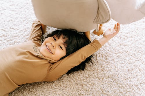 Boy in Brown Long Sleeve Shirt Lying on a Carpet Near a Chair