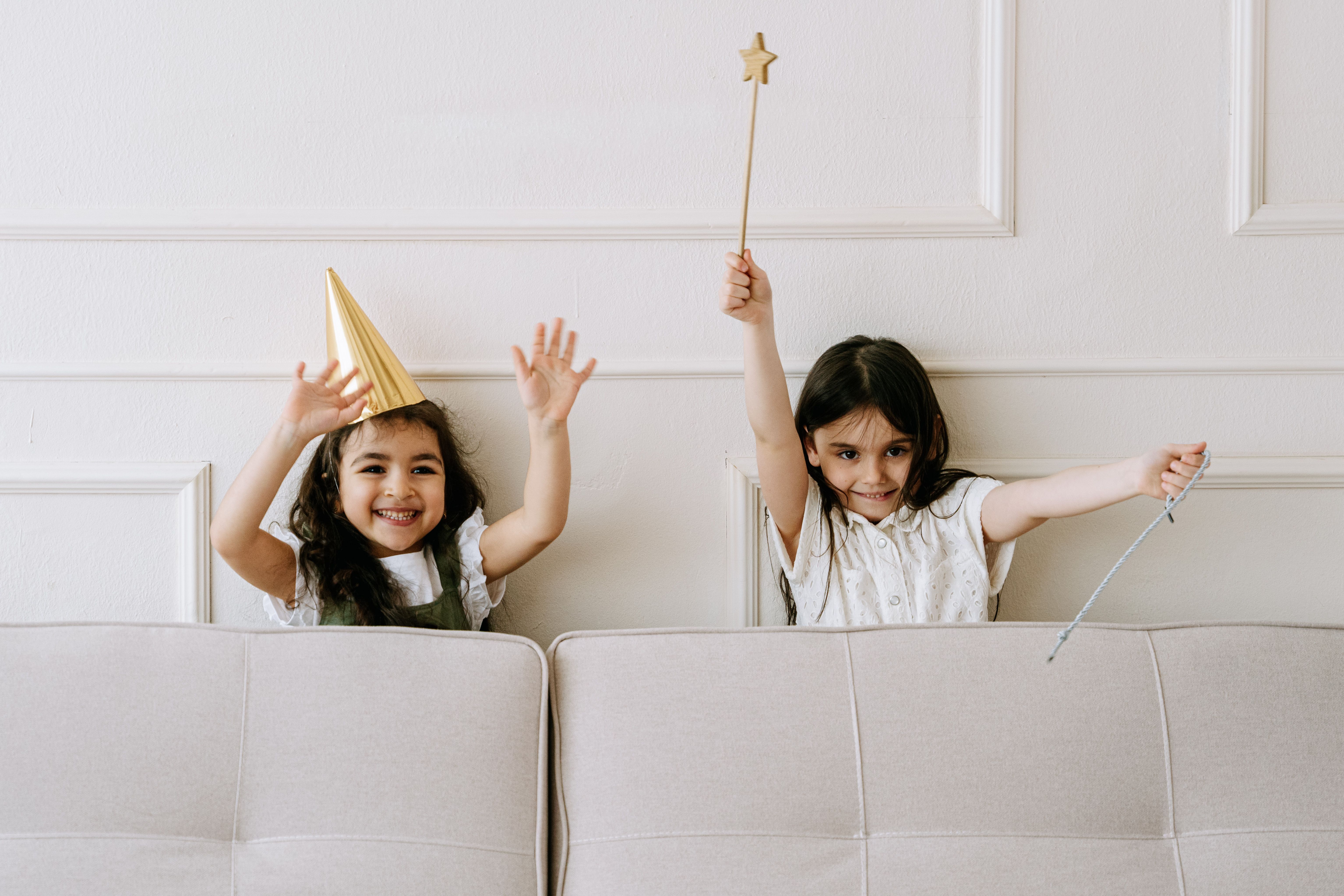 Two smiling children playfully holding magic wands behind a couch indoors.
