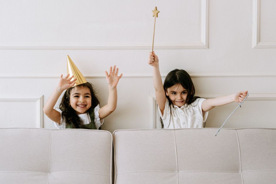 Two smiling children playfully holding magic wands behind a couch indoors.
