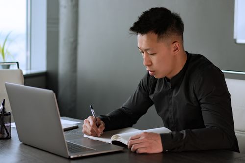 Free A professional man writes notes in a notebook at his desk with a laptop, demonstrating work focus. Stock Photo