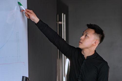 Free Asian man in black long sleeves presenting at a whiteboard in a corporate office setting. Stock Photo