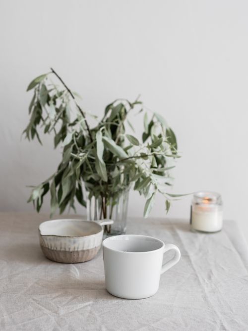 Free A calming still life features a leafy branch, ceramic bowl, mug, and lit candle on a linen tablecloth. Stock Photo