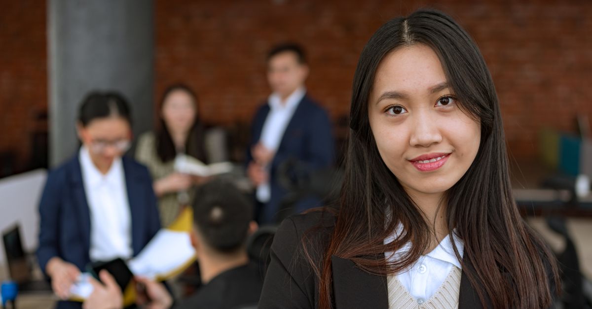Portrait of a confident Asian businesswoman smiling in a modern office with colleagues in the background.