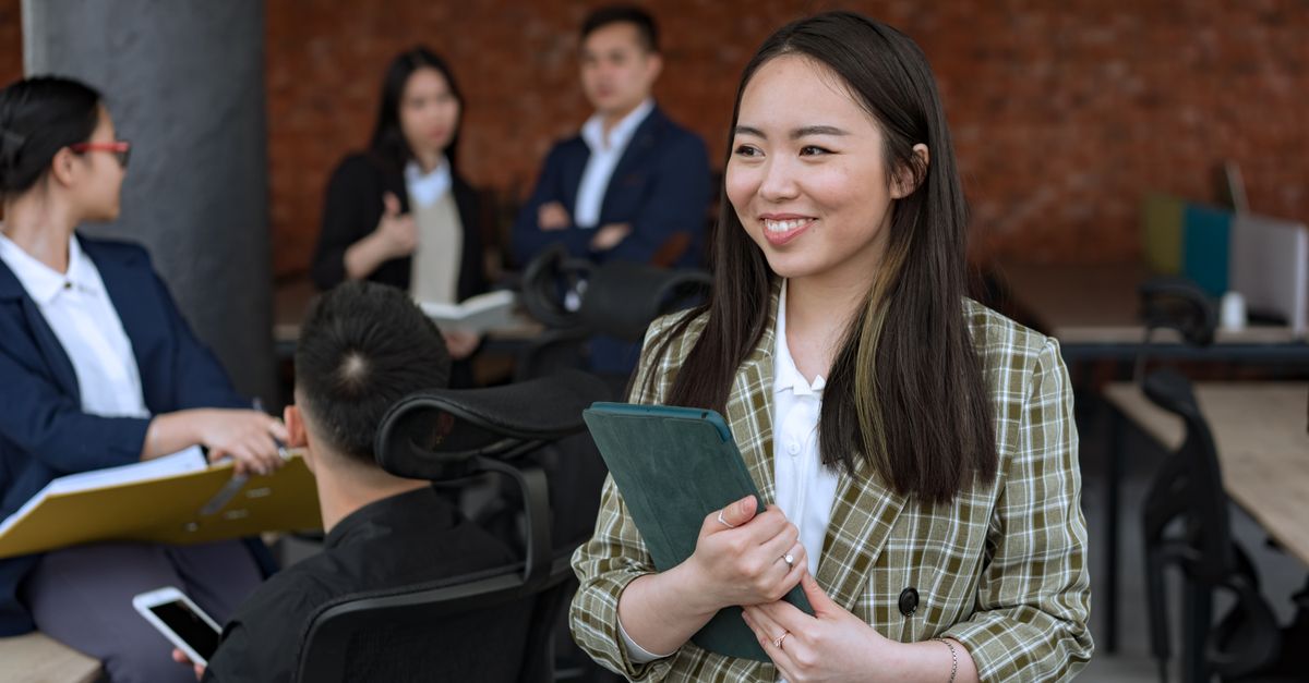 Smiling Asian woman holding tablet in a modern office setting, fostering collaboration.