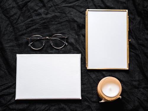 Blank Photo Frame and White Board Beside An Eyeglasses and Candle On Black Textile