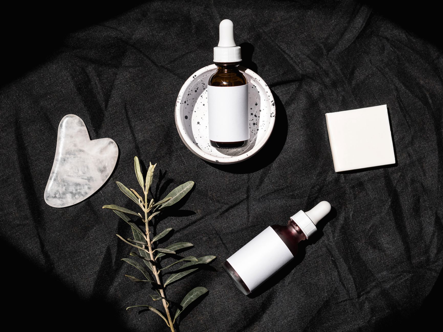 Stylish flat lay of skincare serum bottles with gua sha tool and leaves on a dark textile background.