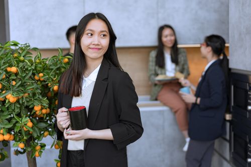 Free Asian woman in a blazer holding a mug, with colleagues in an office kitchen setting. Stock Photo