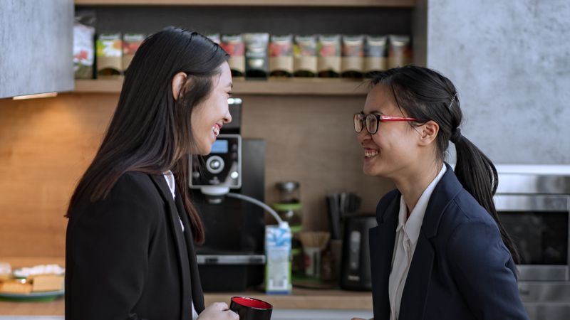 Zwei Kolleginnen in Blazern lachen sich in einer Büroküche an, eine hält einen roten Becher, dahinter eine Kaffeemaschine und ein Regal mit Teepackungen