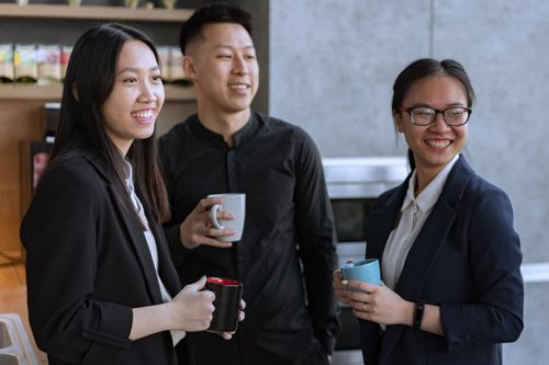 Free Three Asian coworkers enjoying a relaxed coffee break indoors, smiling and holding mugs. Stock Photo