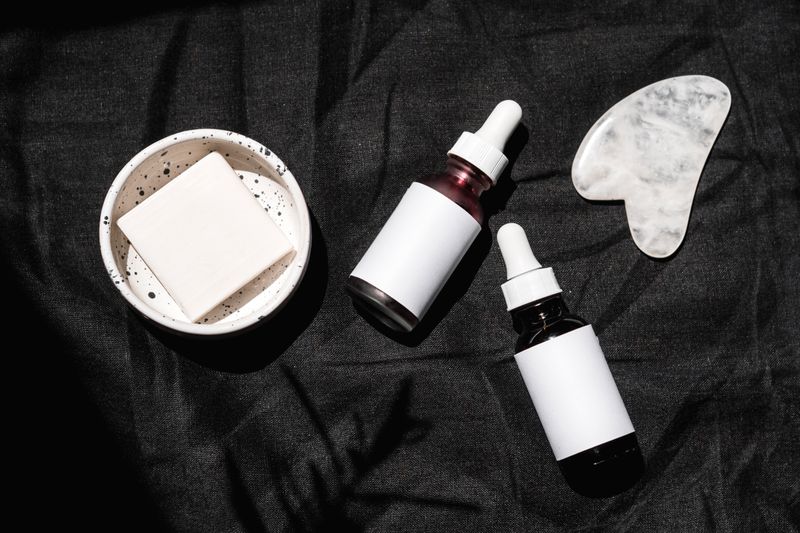 High-angle view of skincare products including serums, gua sha, and bar soap on a dark fabric backdrop.