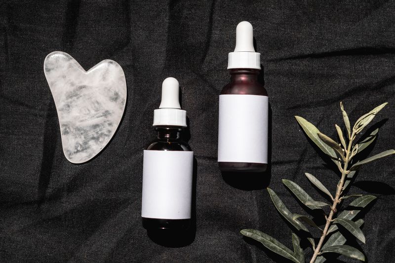 Minimalist flatlay of skincare bottles, gua sha, and a branch on dark fabric background.