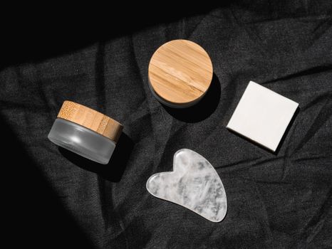 Flat lay of beauty products on black fabric with sunlight, including gua sha stone and containers.