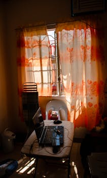 A small room with a laptop on a table, sunlight streaming through patterned curtains.