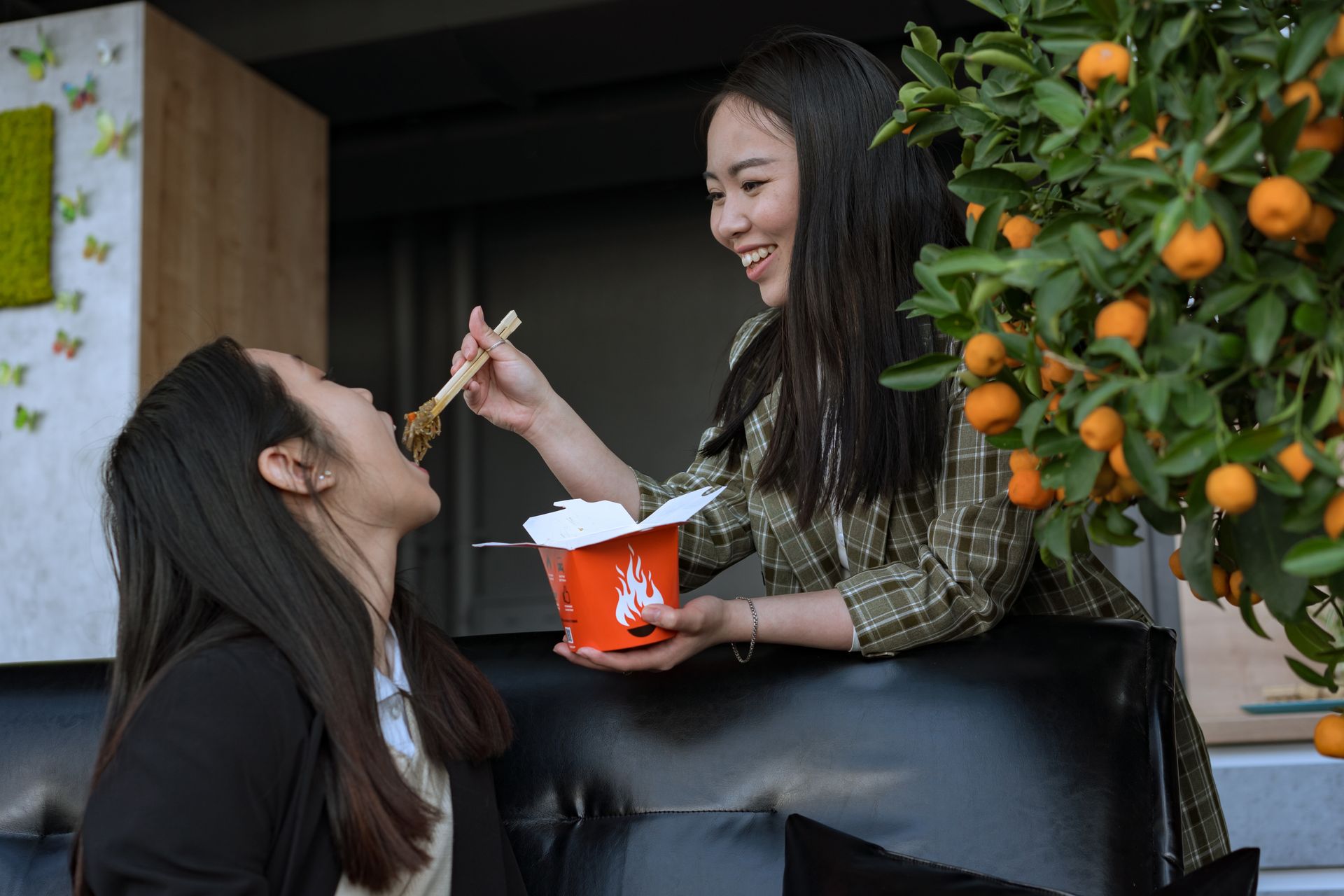 Two women enjoying takeout with chopsticks, sharing joyful moments in an indoor setting.