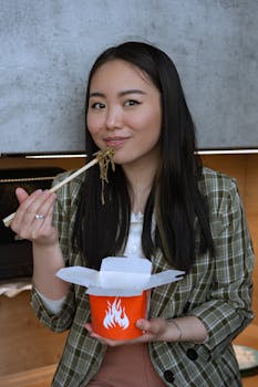 Asian woman savoring noodles from a takeout box, using chopsticks indoors.