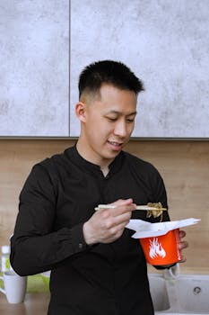 Asian man using chopsticks to eat noodles from a takeout box at home.
